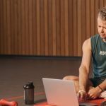 Man sitting on exercise mat with laptop, surrounded by dumbbells, creates a unique fitness workspace.
