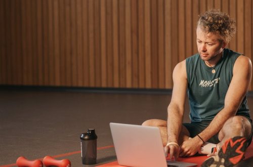 Man sitting on exercise mat with laptop, surrounded by dumbbells, creates a unique fitness workspace.